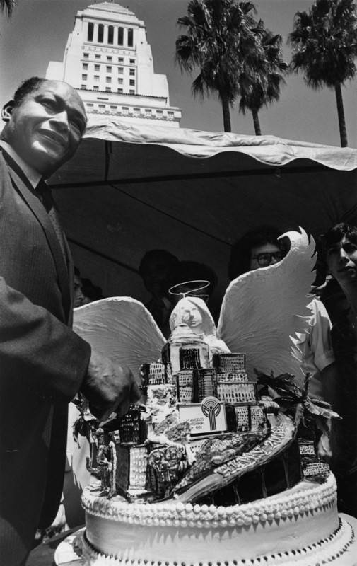 A black and white archival photo of former Los Angeles mayor, Tom Bradley, cutting a cake. The cake is shaped like Los Angeles skyline, with high-rise buildings and palm trees. Behind the cake skyline is an angel with its wings outstretched over the city.