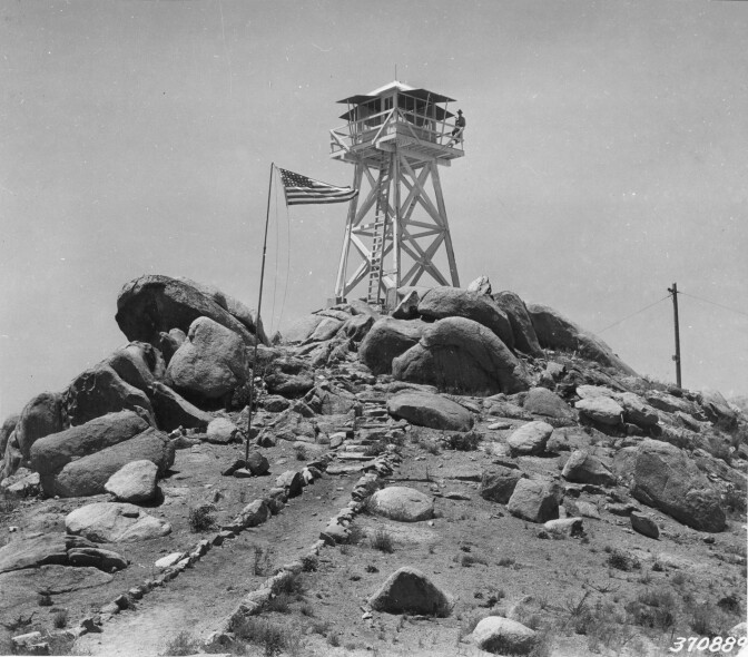 A dirt path leads up a peak of a mountain to a large eight-by-eight-foot wooden fire lookout tower. Boulders sit at the base of the tower, and an American flag sways next to the tower.
