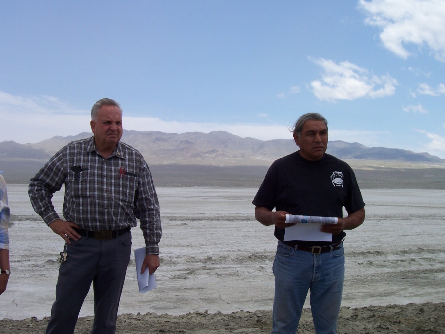 Harry Williams stands in a dry lake bed holding a paper with mountains in the background and another man standing beside him.