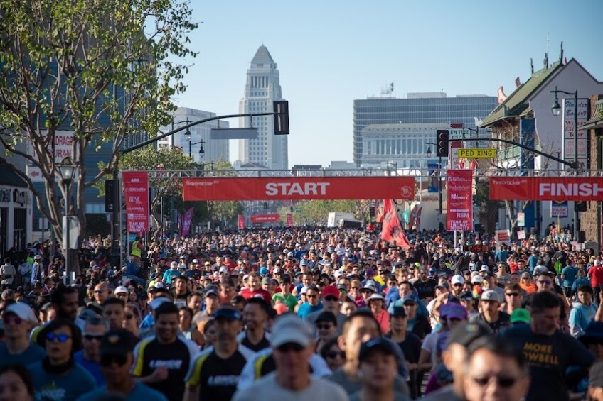 Runners crowd a street in L.A.'s Chinatown as they pass a race starting point, with City Hall in the distance behind them.
