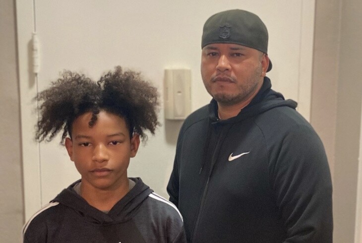 A young boy with black curly hair and a man in a cap wear black hoodies and face the camera.