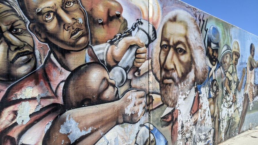 A mural showing Frederick Douglass in the middle, flanked by an African American man holding an African American child on one side and a Black soldiers and slaves on the other side.