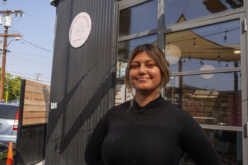 Justine Hernandez, wearing a black long-sleeved turtleneck shirt stands in front of the Just What I Kneaded bakery storefront. The building is painted black with large wall windows. A small circular sign that reads, "Just What I Kneaded" along with the bakery logo of dough being rolled is posted on the wall.