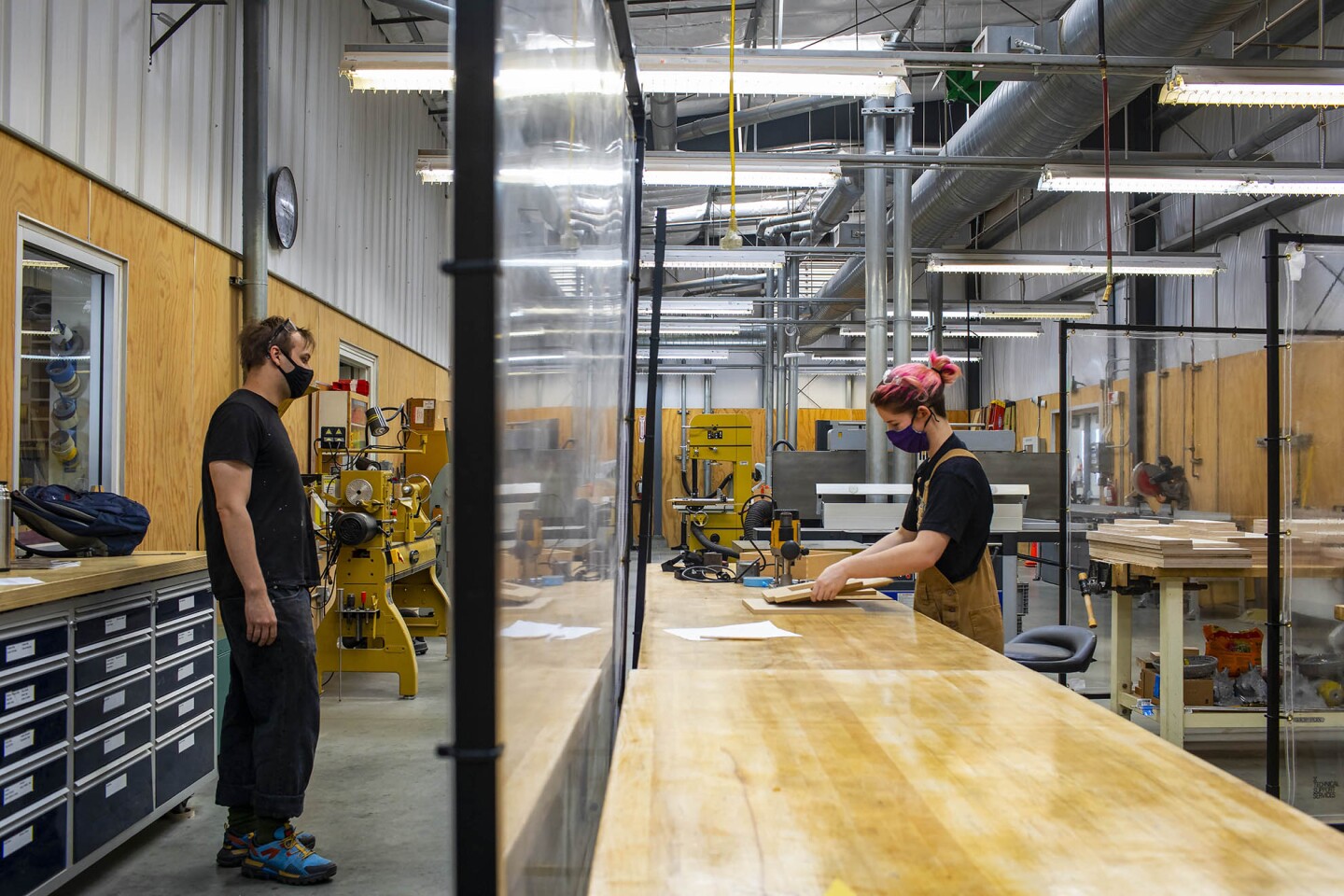 A man (left) and a woman (right) in masks work in a woodshop with coronavirus safety regulations in place such as clear plastic barriers between stations.