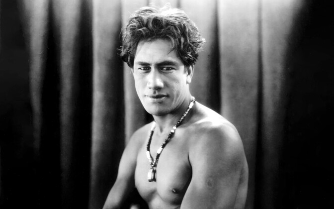 A black and white portrait photo of Duke Kahanamoku. He stands shirtless, only wearing a beaded necklace around his neck. He has dark hair and is turned to the camera with a slight smile.
