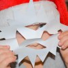 A small child wearing a red Santa hat holds a paper snowflake in front of the face like a mask.
