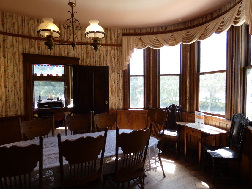 An oval-shaped dining room with floral wallpaper and curved glass windows. A the center is a wooden oval dining table with wooden chairs. 