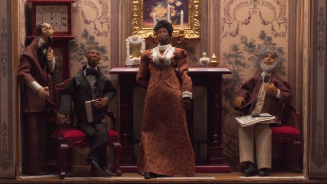 A miniature diorama depicts a small, clay figure of Madam C.J. Walker standing in a burnt orange dress with an ornate collar. Behind her is a fireplace. Around her, three men sit around.