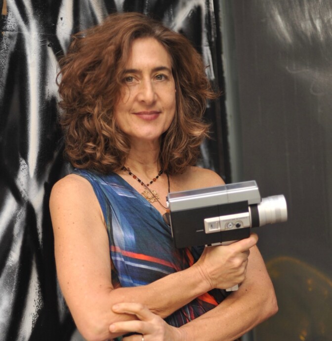A woman in a dress holds a small video camera in her hand while leaning on a wall