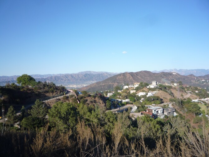 A scenic landscape photo of rolling mountains. A road carves through certain parts and several houses and buildings are sparsely dotted on the right side of the photo.