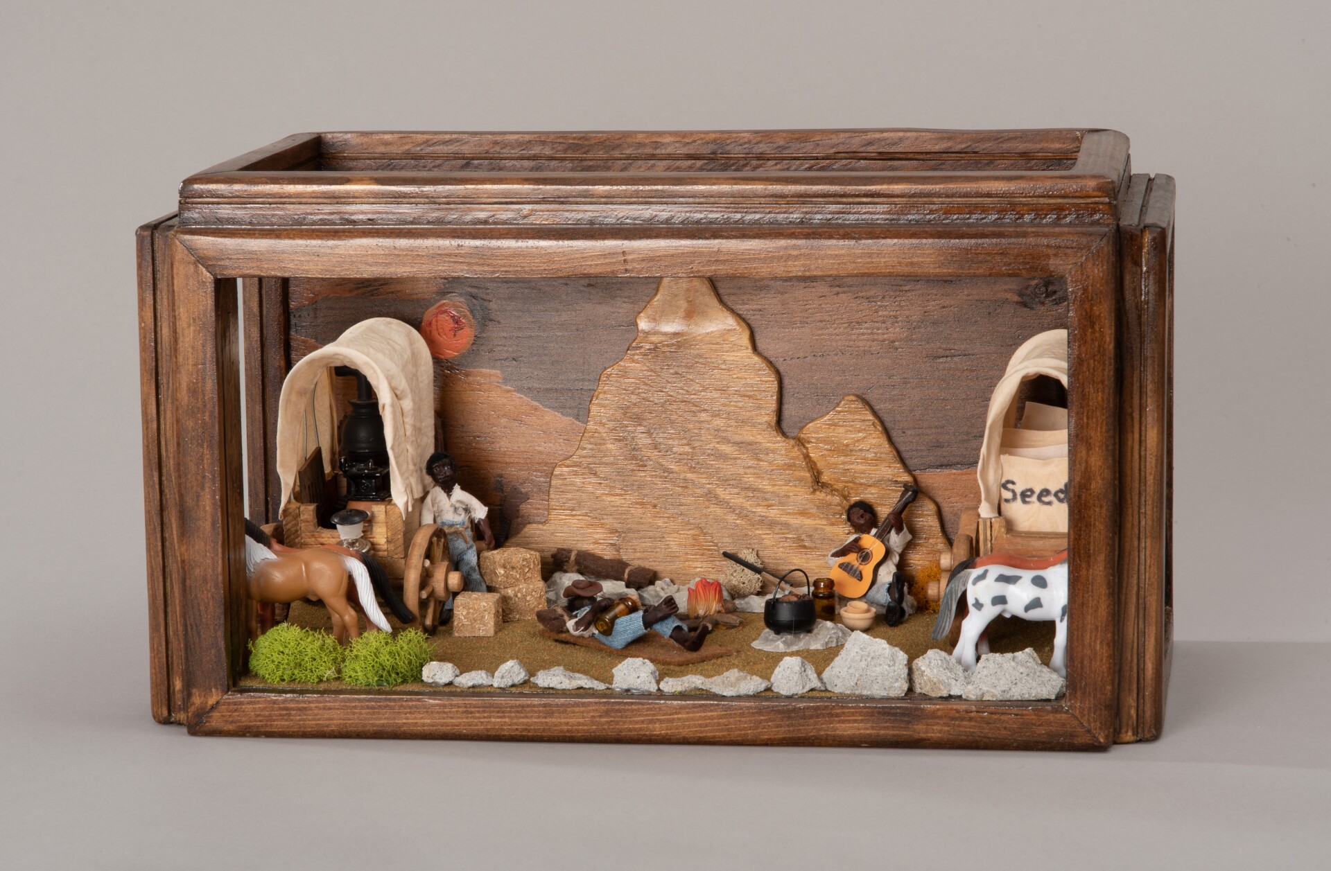 A miniature diorama is contained inside a shadowbox with wooden frames. Inside the shadowbox depicts a scene of Exodusters resting in a wild West, desert landscape. Two Exodusters are gathered around a fire. Around them are carriages, various supplies and a horse.