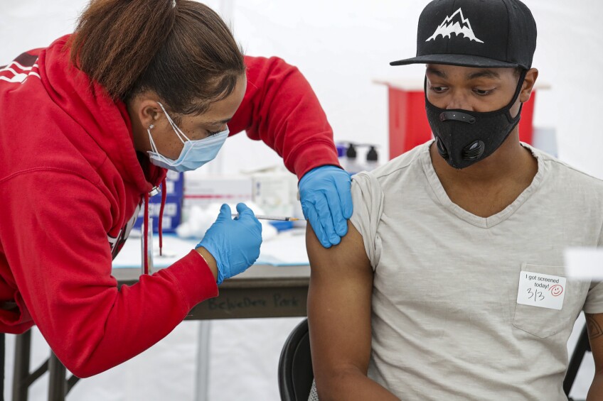 A person in a red sweatshirt administers COVID-19 vaccine to a young Black man. Both are wearing face masks.