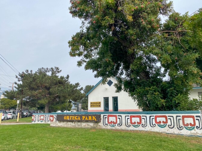 A welcome sign reads, "Ortega Park." The letters are painted yellow on a dark brown placard. The sign is fixed on a low wall that is painted with decorative symbols, using red and green paint over a white background. The symbols appear pre-Colonial in nature.