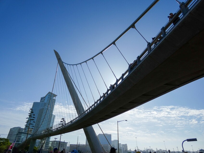 A winding concrete footbridge with a steel frame and wires that connect it to the bridge stands against a bright blue sky. Tall, city buildings can be seen off to the left. 