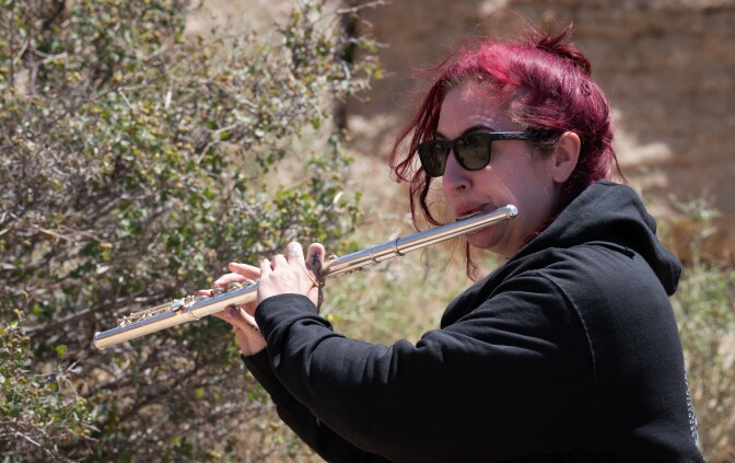 Christine Tavolacci plays the flute, holding the mouthpiece up to her lips as she blows into the instrument, her fingers hovering over the keys. Christine has bright red/maroon hair that is tied in a bun. She is wearing shades and a black zip-up hoodie.