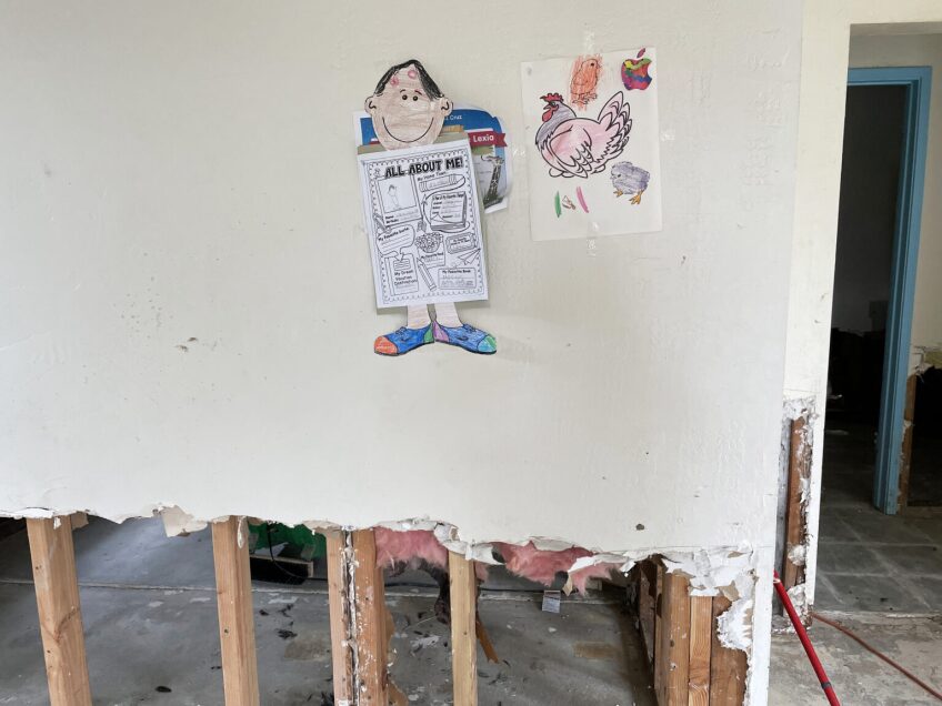 Children's coloring pages and other papers from school hang on a beige wall whose drywall has been stripped away several feet above the floor, exposing the wood studs underneath.