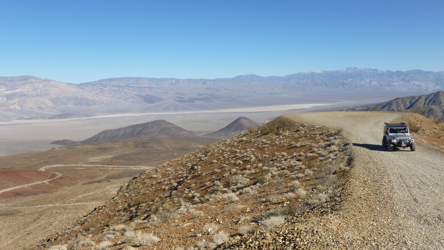 A silver jeep sits on a dirt path with a wide view of Death Valley behind it.