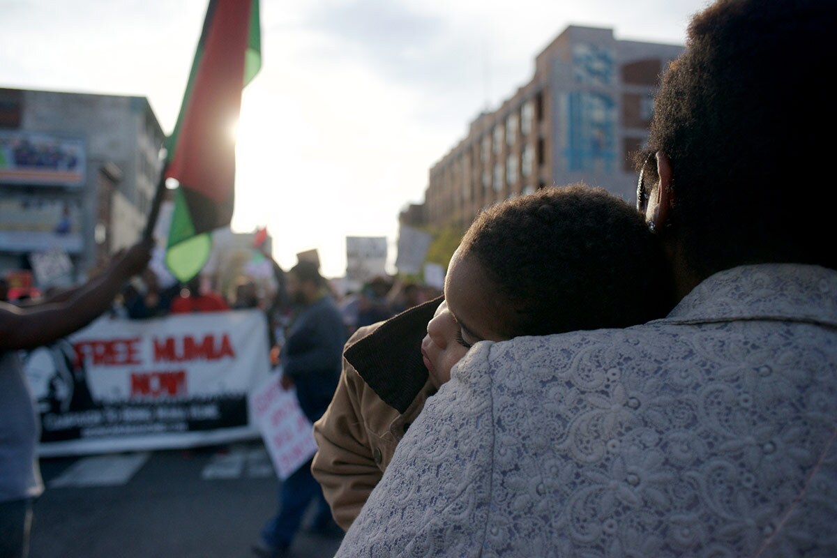 A parent holds a small child on their chest at a protest while the sun's rays shine on them.