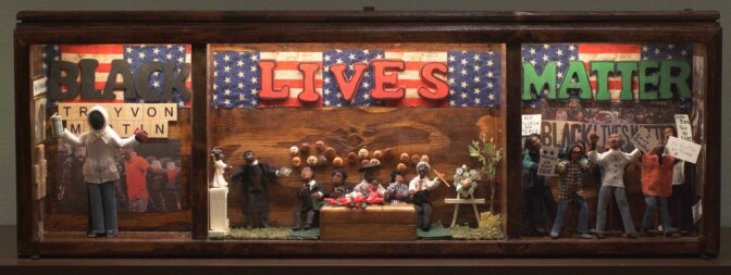 A three panel shadowbox diorama with wooden frames pays tribute to the Black Lives Matter movement. The first panel depicts Trayvon Martin with his hands up, holding a can of Arizona tea in one hand and Skittles in the other. Behind him are photos of protest and the word, "Black" cut out with black construction paper. The center panel depicts people gathered around a casket and the word, "Lives" above them cut out with red paper. The last panel depicts clay figures posed in protest, holding signs and images of protest behind them. Above the scene is the word, "Matter" cut out in green construction paper.