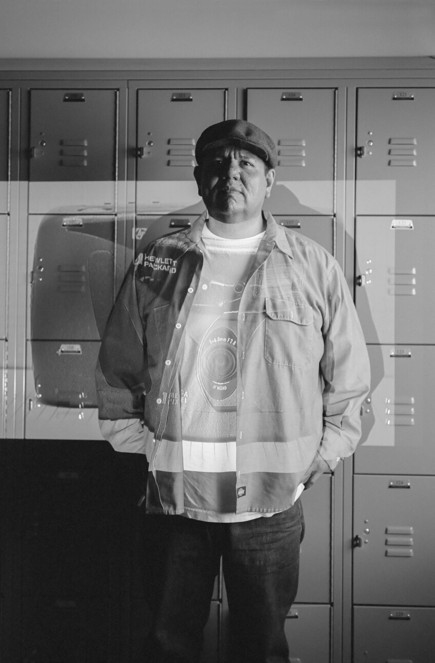 A black and white photo of a man standing with his hands in his pockets and in front of a wall of cube lockers. Layered over him is a transparent image of a camera.
