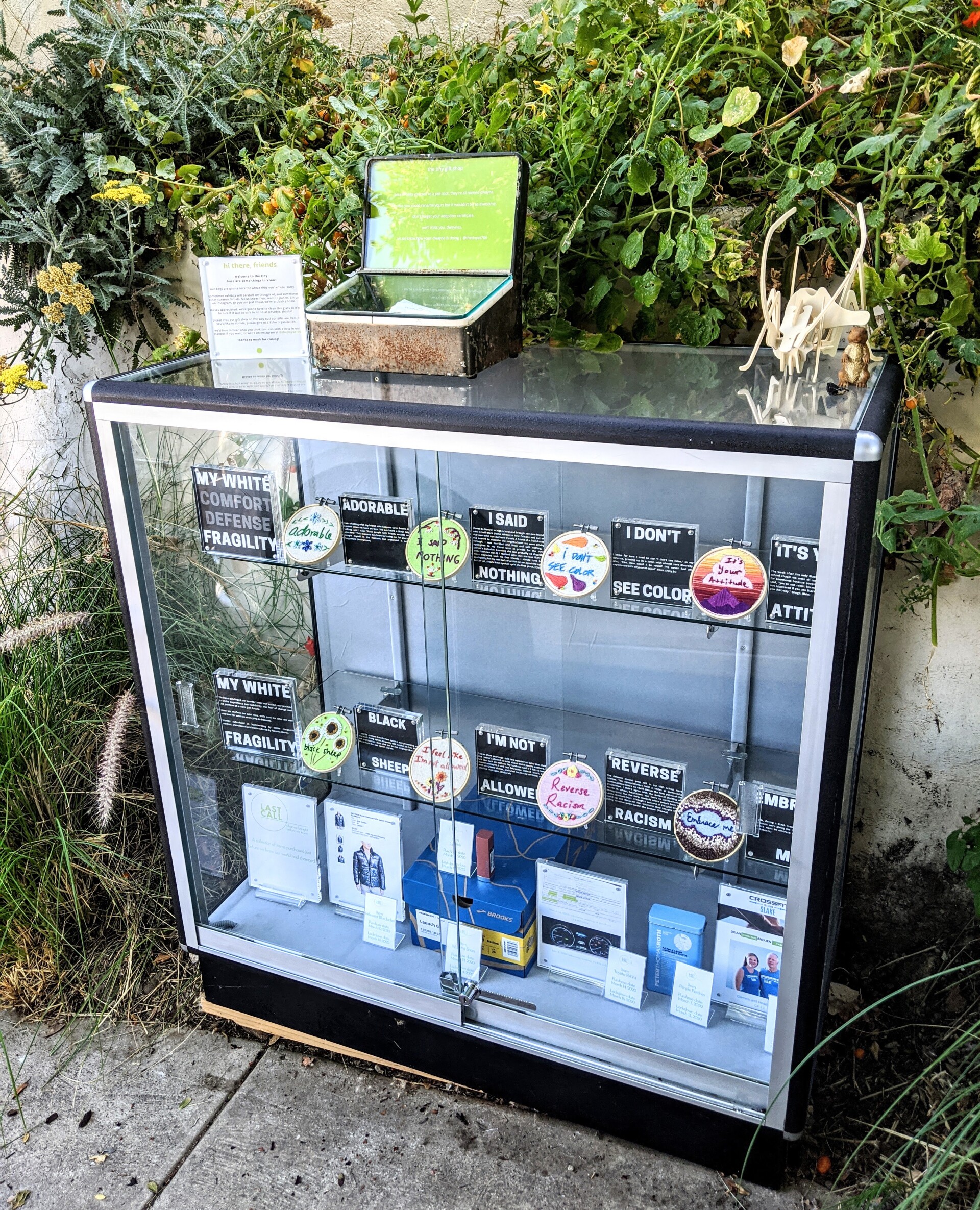 Eight embroidery designs of various colors and phrases centered around the theme of white fragility sit behind a three-shelf glass display. Behind it is a concrete wall covered in thick green foliage.
