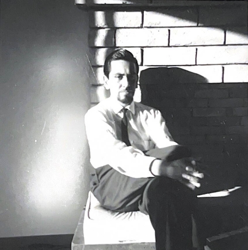An archival black and white photo of J. Sergio O’Cadiz Moctezuma sitting on the mantle of a fireplace. His leg is crossed over the other and his hands are folded over his knee. He's wearing black slacks and a black tie and looks directly at the camera.