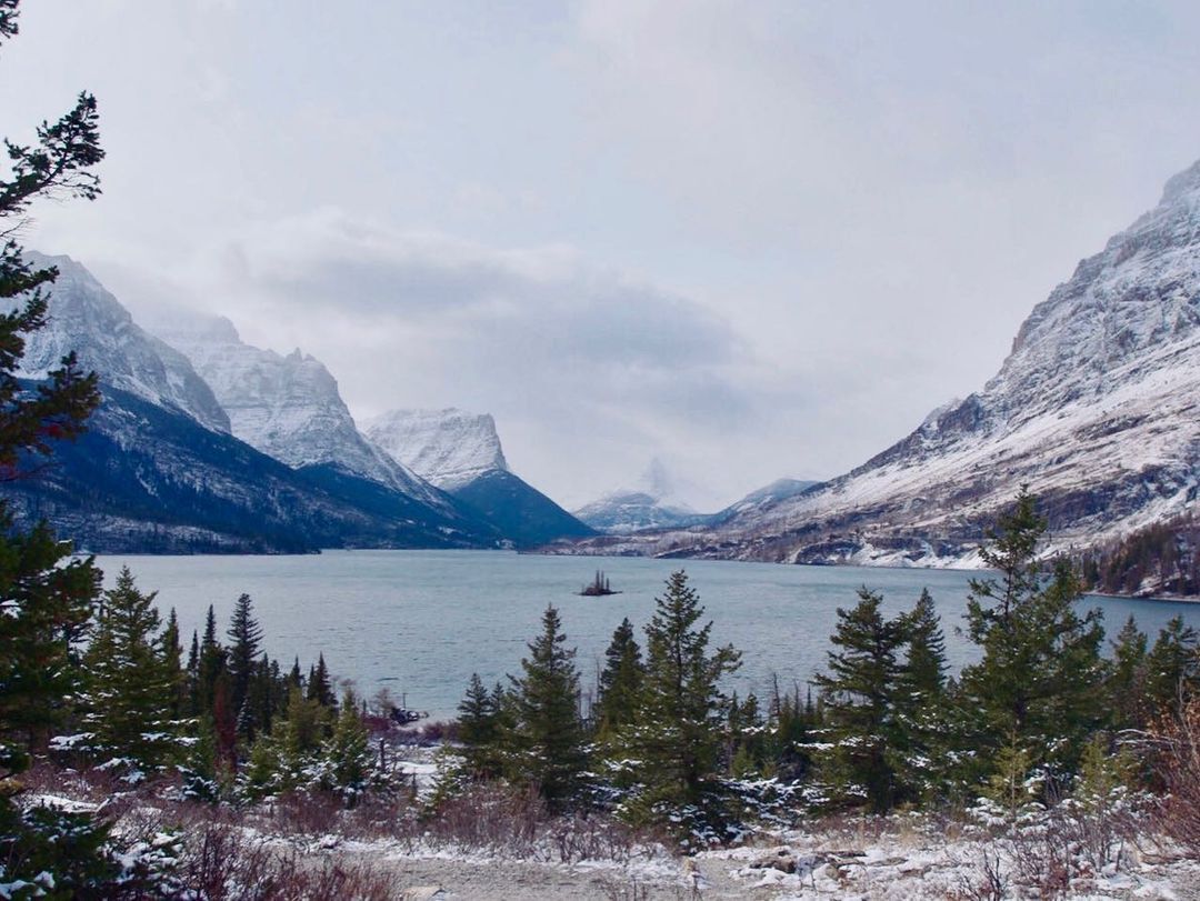 A scenic landscape captures ice-capped mountains surrounding a clear, blue lake. At the foreground are green pine trees dusted with white, powdery snow.
