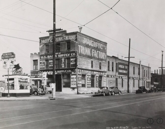 View of brick buildings housing businesses and a General gas station on Aliso Street near Old Chinatown, Los Angeles, with automobiles on the street. The businesses include the Pacific Trunk Factory, U.S. Bag & Supply Co. and American Toy Manufacturing Co.