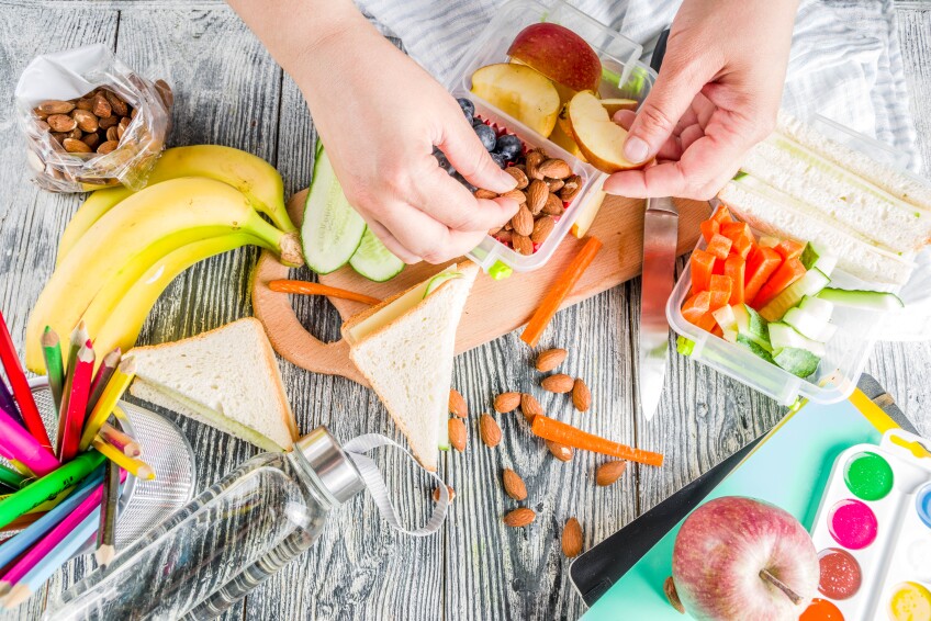 Close up of a person's hands making snack boxes with fruit, nuts and sandwiches.