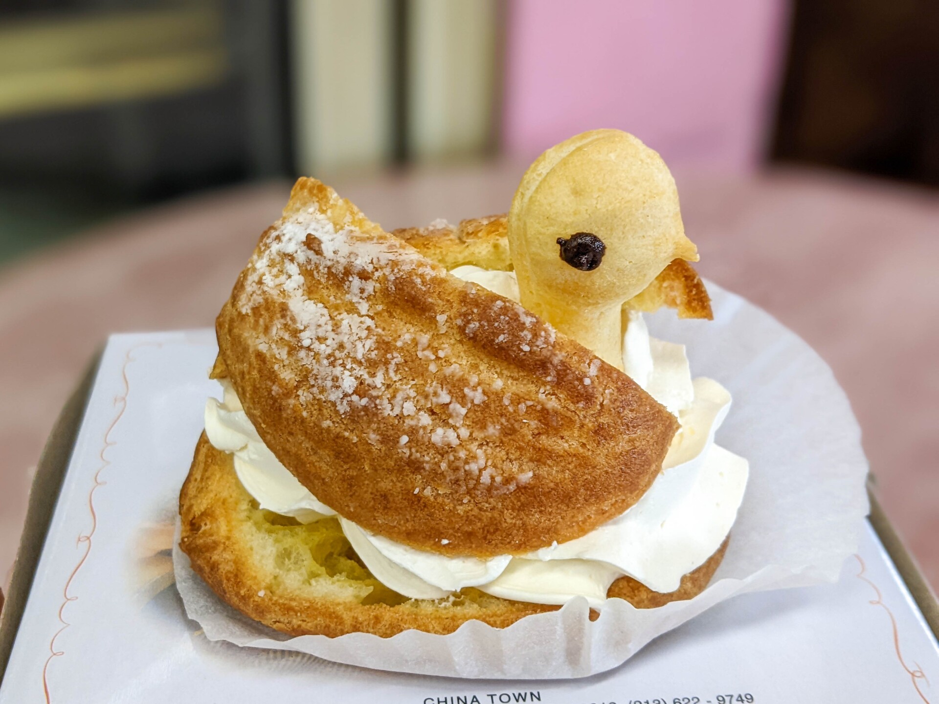 A close-up photo of a golden yellow pastry in the shape of a swan. The pastry is filled with a white cream and the whole thing sits atop a wax paper cupcake liner.