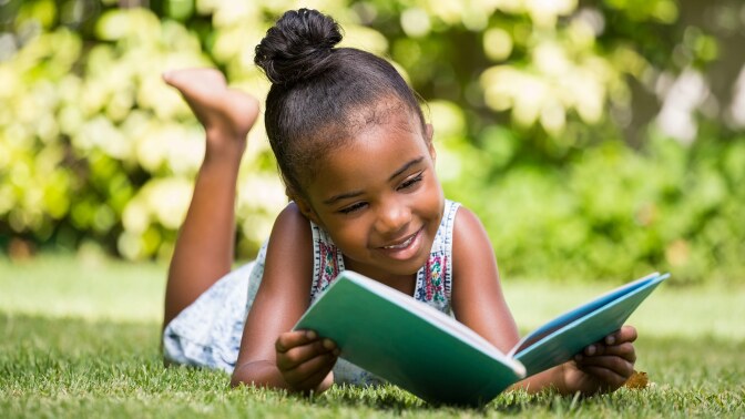 A small child smiles as she reads a book in a grassy place.