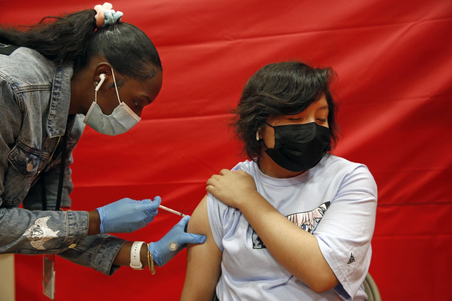 A young man in long black hair gets a vaccine from a young Black woman wearing a ponytail.