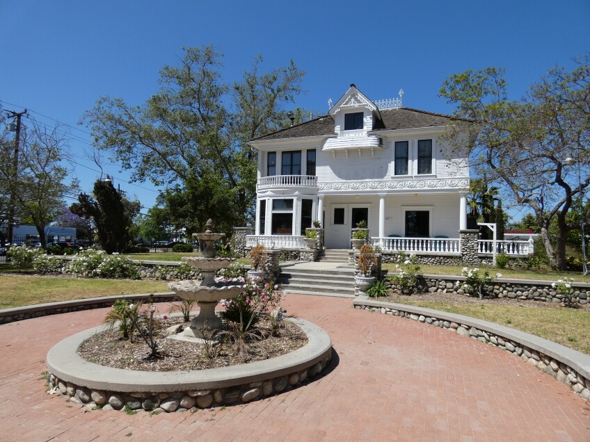 A white Victorian-style house with a large porch sits in front of a circular brick walkway with a fountain centered in the middle. The fountain is dry and surrounded by plants.  