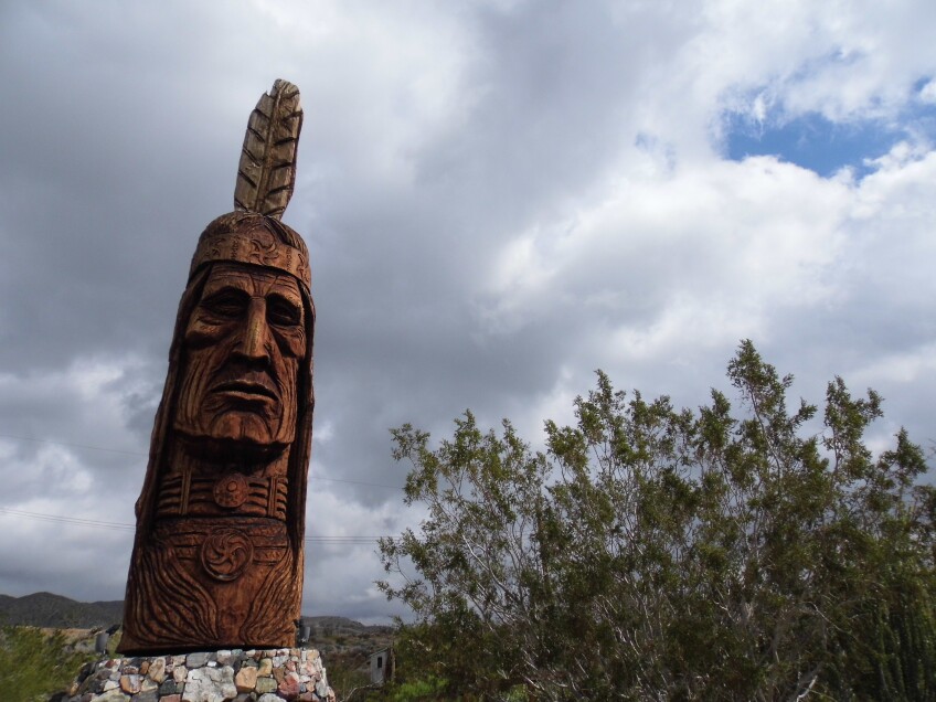 Waokiye, a wooden carving of a Native American head from a sequoia log. The carving stands against a gray, cloudy sky. Rolling mountains can be seen off in the distance. 