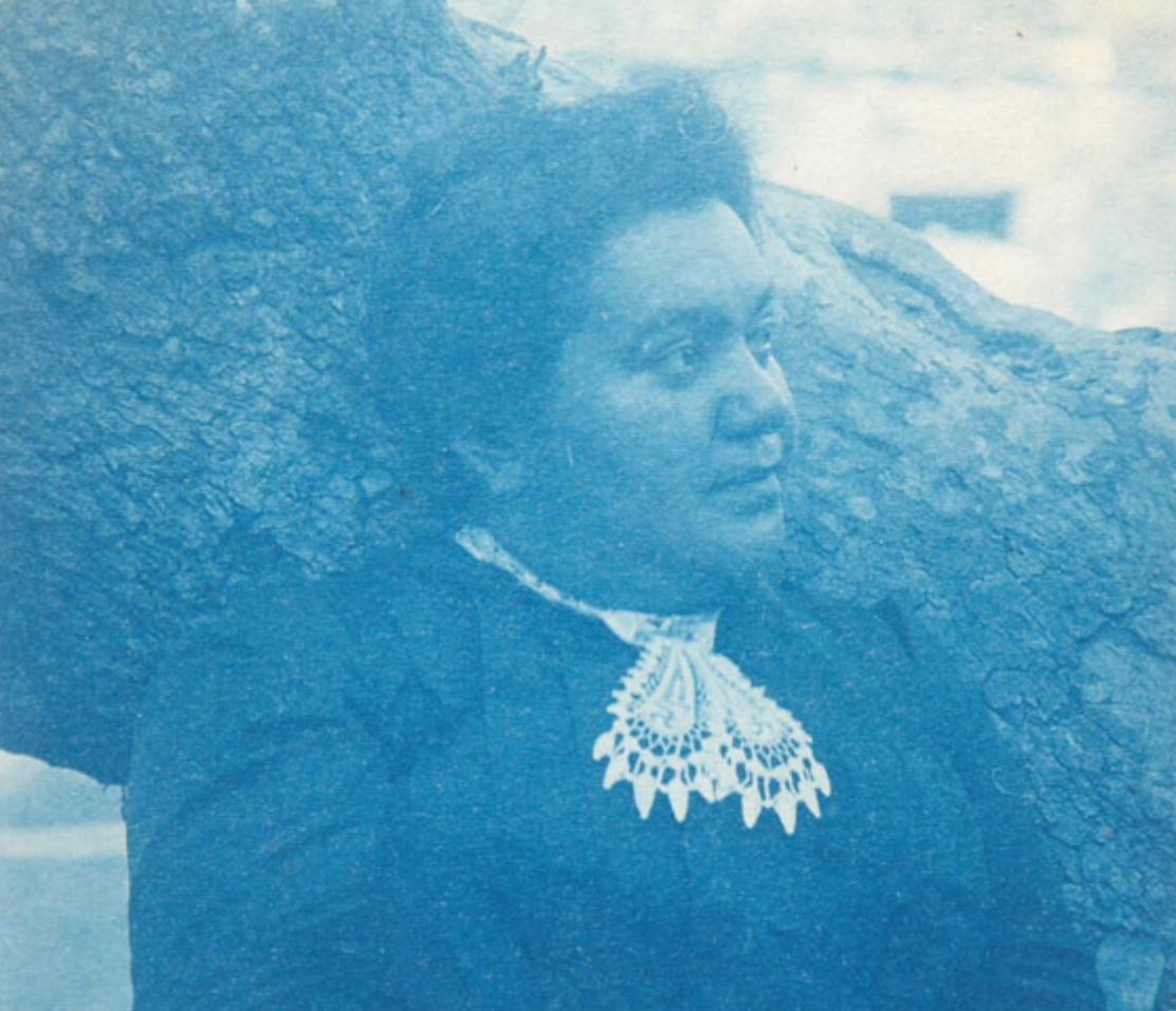 A blue-tinted portrait of Manuela C. García. She's wearing a long sleeved blouse with a lace neck accessory. She looks to her left and stands in front of a thick tree branch.