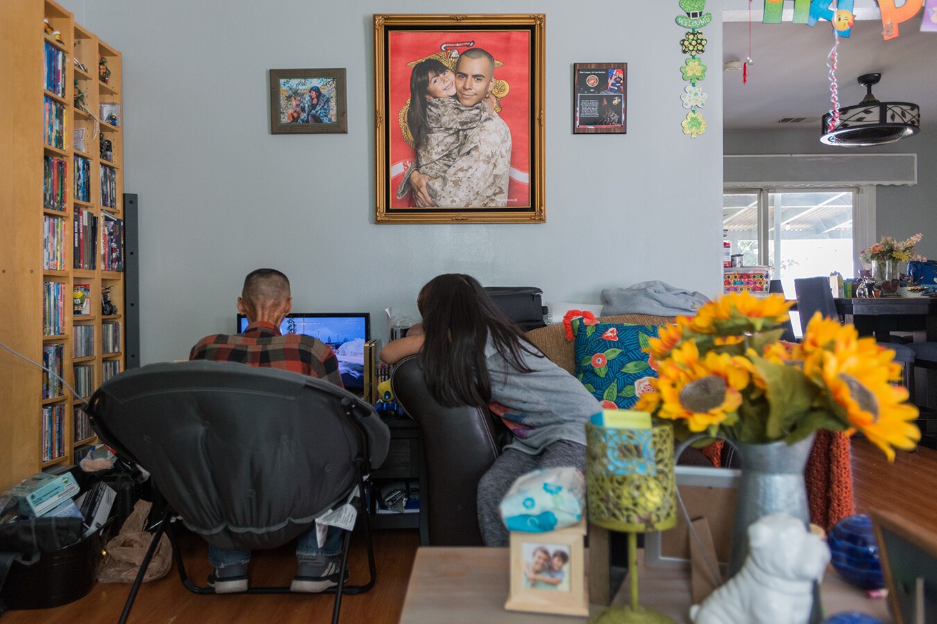 Rhianna watches her dad play a video game on a computer. Their backs are turned to the camera. In frame, you can see their home which is filled with sunflowers in a vase, photos of Rhianna and her dad, a shelf full of DVDs and vibrant throw pillows.