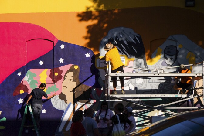 Painters work on a bright and vibrant mural. Two painters are painting on a scaffold positioned by the wall. Another painter is standing on a green ladder. The mural wall is partially shaded by a tree, creating a dynamic look to the wall.