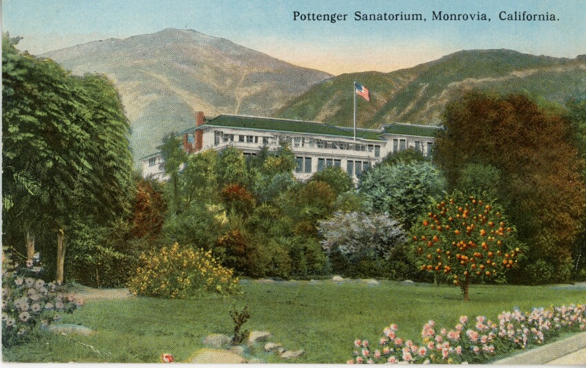 A hand-colored postcard of a large, white, colonial-style building with a green tiled roof stands behind a lush landscape of flower beds, a green lawn and many varieties of trees, with mountains looming just behind. An American flag waves at the top of a flagpole above the roof.