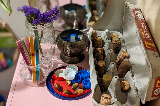 Loose parts including popsicle sticks, bottle caps, pieces or cork, empty egg cartons, and more are arranged on a pink table.