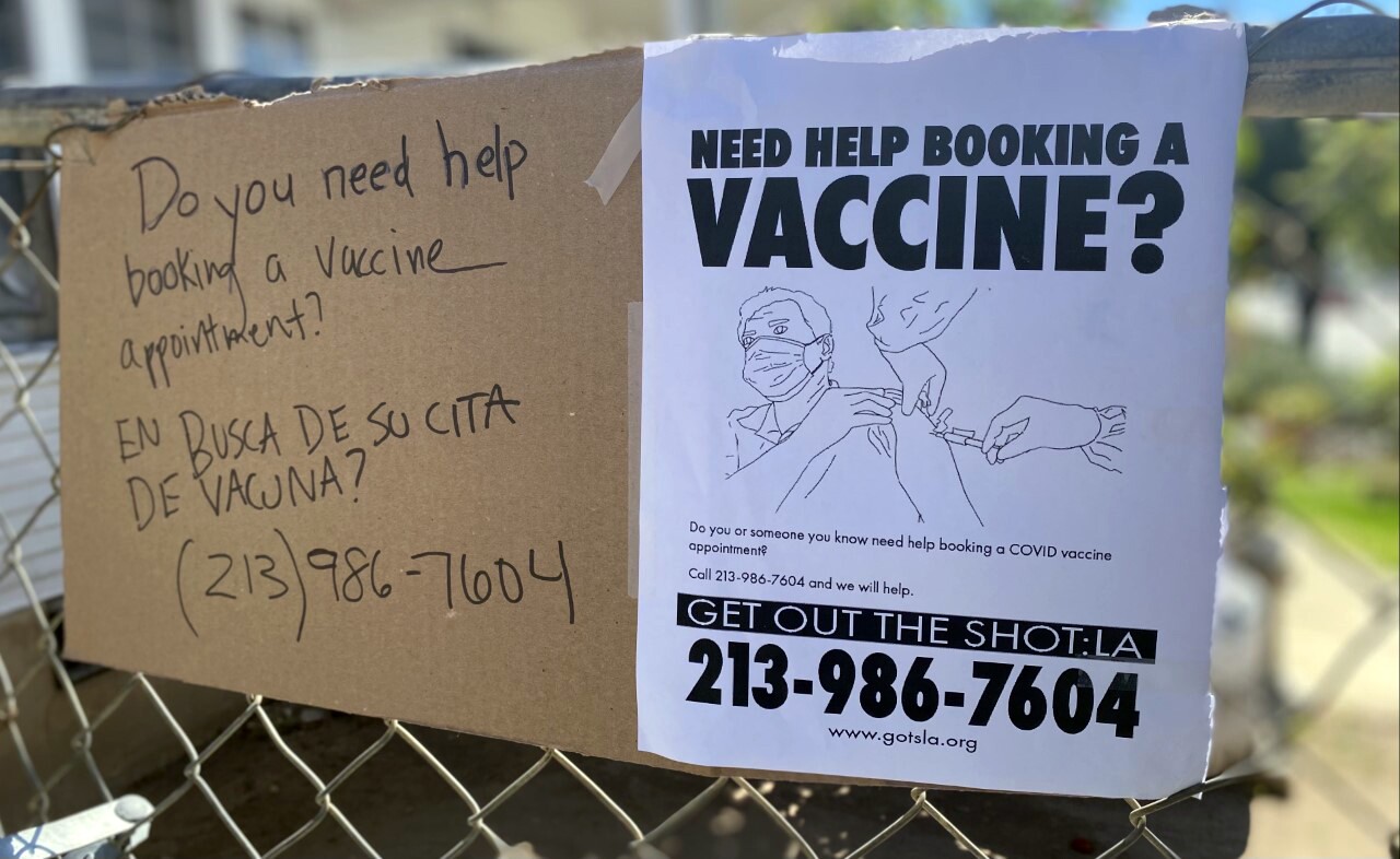 A cardboard and paper sign has a picture of a person being vaccinated and reads "Need help booking a vaccine appointment?" and lists the number 213-986-7604 and the website www.gotsla.org.