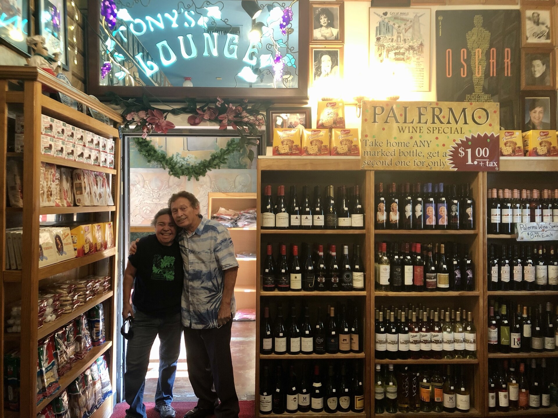 Dora Herrera and Tony Fanara stand in a doorway. Above them is a mirror neon sign that reads "Tony's Lounge" in teal blue and decorative purple grape designs. To their right is a shelf displaying various wines. To their left is a another shelf unit containing different snacks and pastries. 