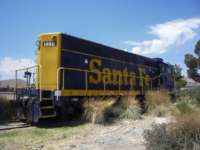 An antique railroad car sits on a patch of dirt outside. The train car is a dark, navy blue with yellow detailing and trim as well as the word, "Santa Fe" written across in the same yellow color.