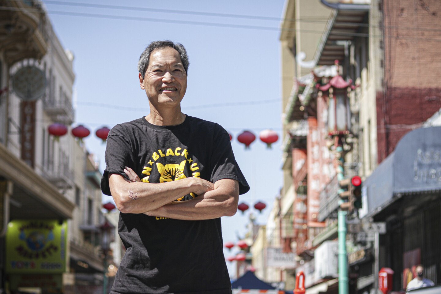 A wrinkled Asian American man in a black t-shirt grins as he crosses his arms across his chest. He stands in Chinatown, San Francisco.