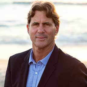 Man in blazer and blue collared shirt, standing in front of a beach sunset.