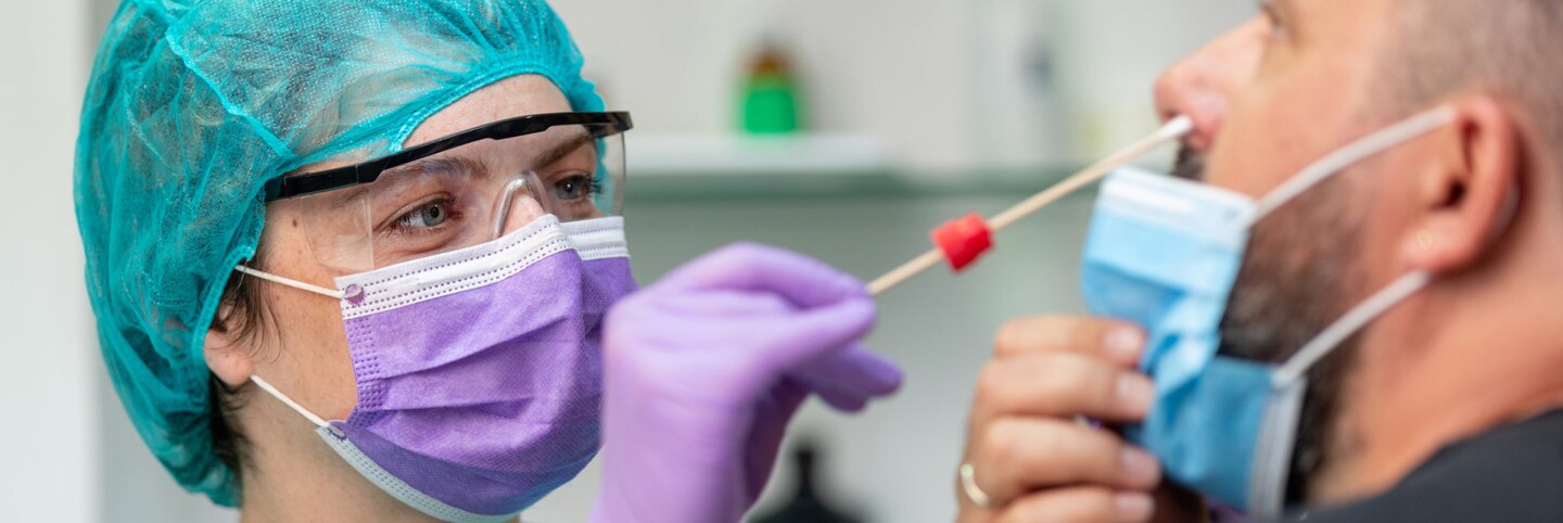 A female doctor in protective workwear does a nose swab COVID-19 test on middle aged man wearing a protective face mask. | iStock