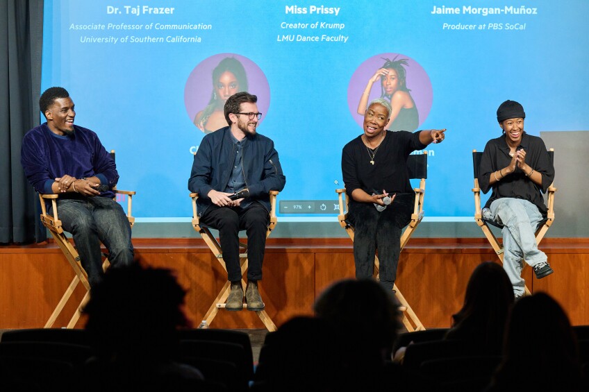A panel of four people sit in director's chairs with amused expressions, and the third person from the left points to the crowd.
