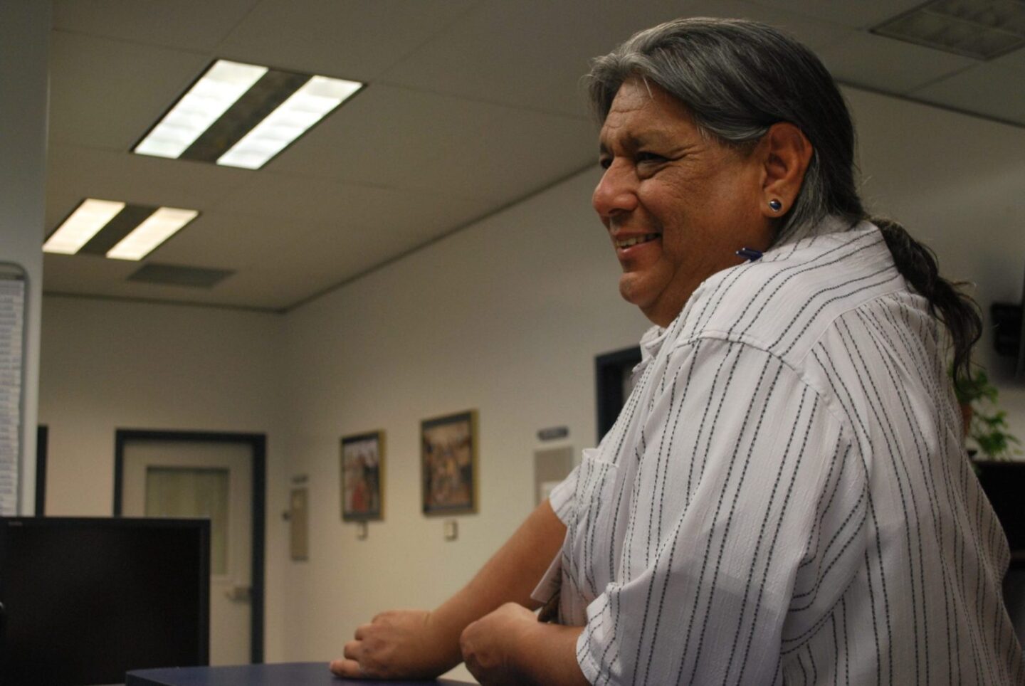 Side photo of an Indigenous man who is smiling inside an office. He is wearing his long hair in a ponytail and wears a striped button down shirt.