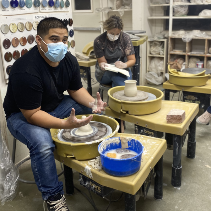 A man sits by a potter's wheel explaining ceramics.