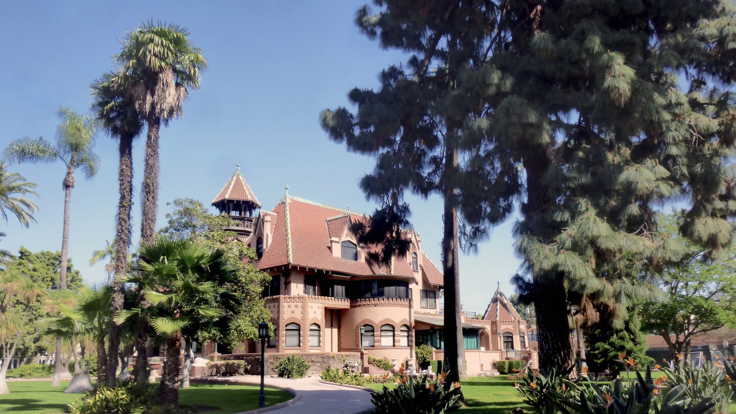 The Doheny mansion, an red-orange brick building of French Chateauesque style architecture, mixed with Gothic, Moorish and California Mission elements stands among a grove of various trees. A paved pathway winds up to the mansion. Above the mansion is a bright blue sky.