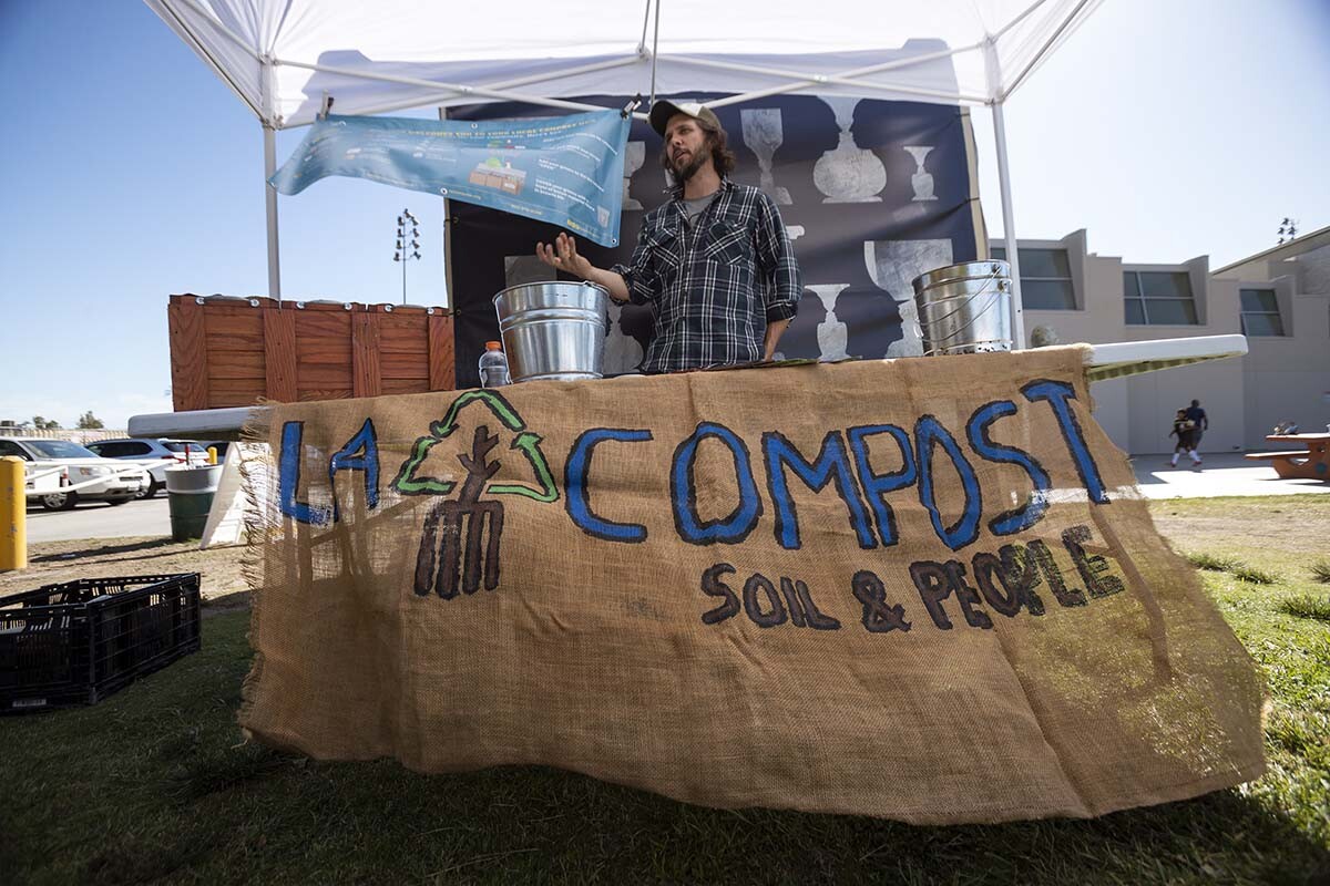 A man stands behind a stall with the words "L.A. Compost" while rehearsals of “ALL AGAIN” goes on. The event was organized by Carolyn Pennypacker Riggs and Annie Gimas | Courtesy Department of Cultural Affairs, City of Los Angeles
